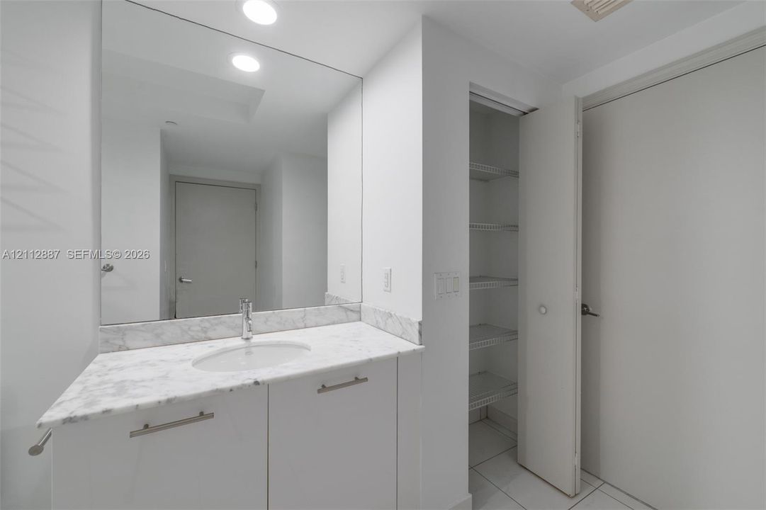 Vanity featuring a stone-surface countertop with an integrated oval sink, full-width wall mirror, white cabinetry with brushed metal hardware, and a built-in linen closet with wire shelving