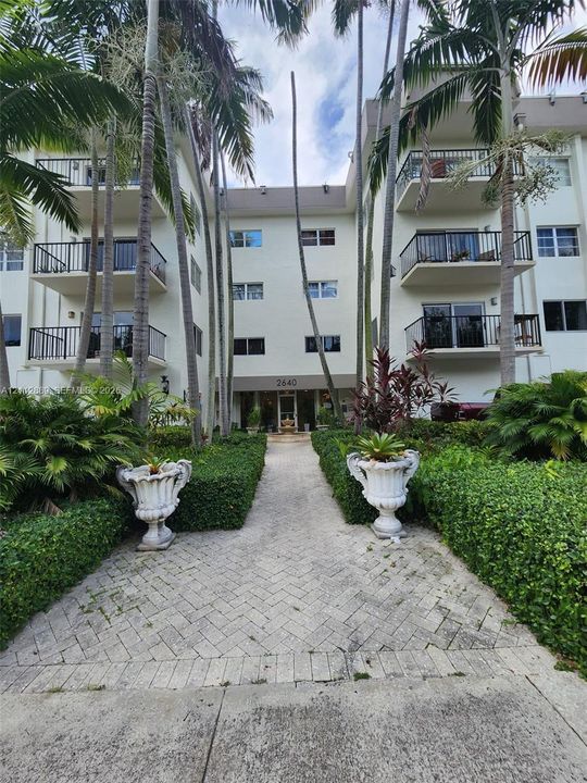 Multi-story building exterior featuring a herringbone-patterned stone walkway, manicured hedges, mature palm trees, and private balconies with black metal railings