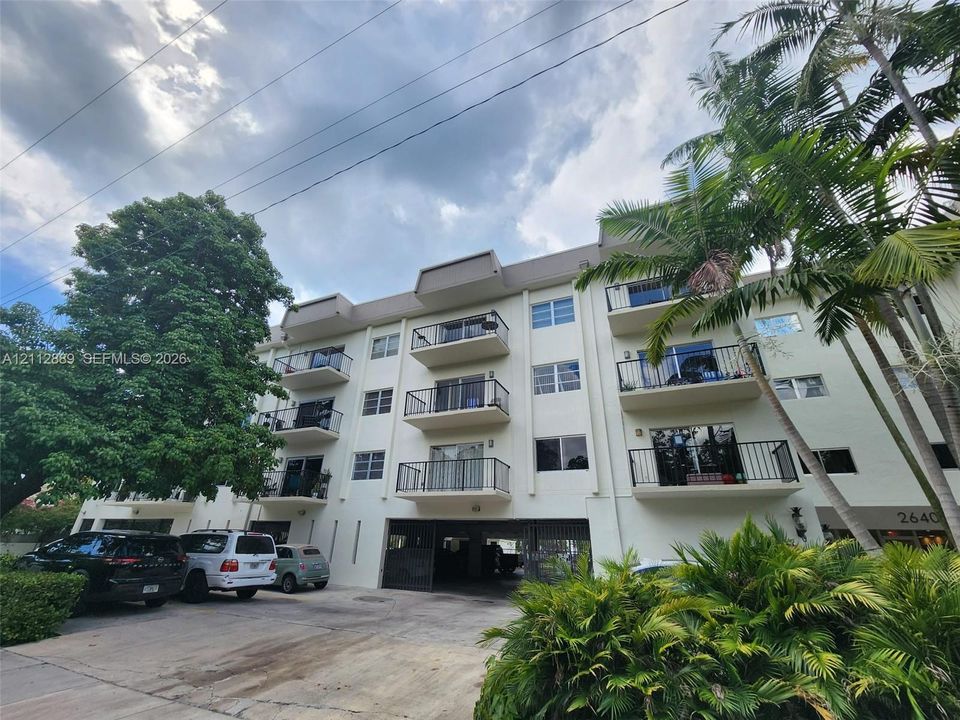 Multi-story building exterior featuring concrete construction, private balconies with metal railings, and a covered ground-level parking area