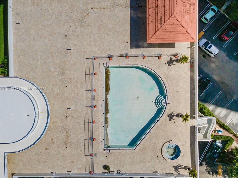Aerial view of a sprawling pool deck featuring stone-tiled hardscaping, an in-ground swimming pool, a separate circular spa, and a covered pavilion structure