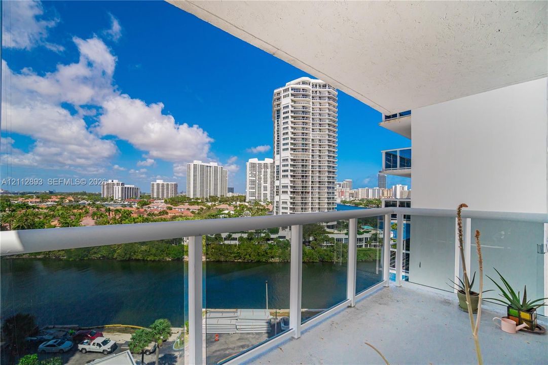 Covered balcony featuring glass panel railings and a tubular white metal handrail, offering exposure to the surrounding waterway and high-rise architecture