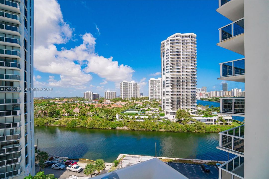 High-rise balcony featuring glass railings, overlooking a waterway, mature landscaping, and an expansive cityscape