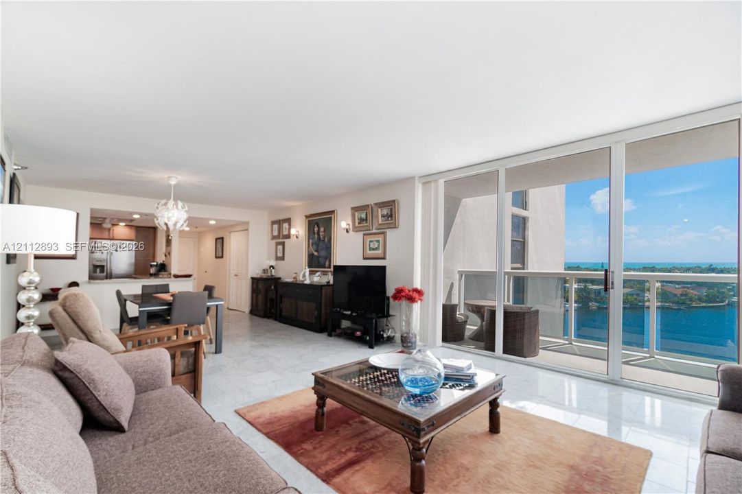 Open-concept living area featuring light-toned tile flooring, floor-to-ceiling sliding glass doors, a balcony with a glass railing, and custom overhead lighting fixtures