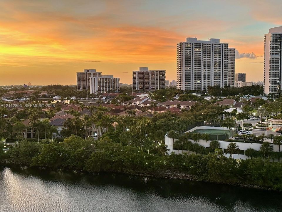 Elevated perspective featuring high-rise architecture, a waterway, mature tropical landscaping, and a tennis court
