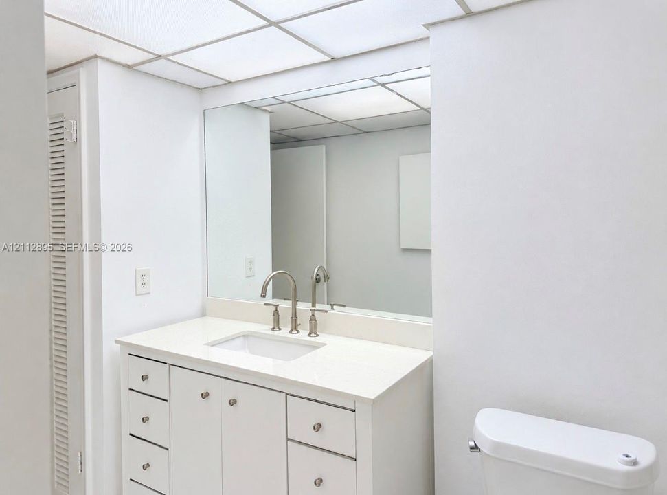 White vanity cabinet with integrated countertop, undermount rectangular sink, bridge-style faucet, large wall-mounted mirror, and drop ceiling grid lighting