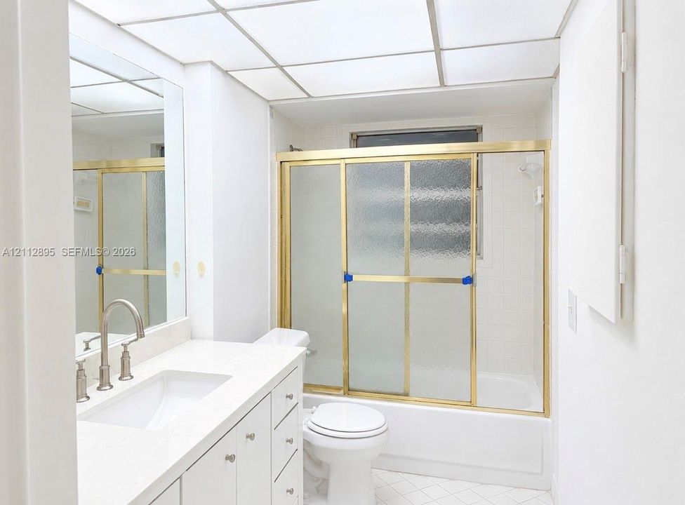 Bathroom featuring a white vanity with a solid-surface countertop, an undermount sink, a drop-in tub with sliding glass doors, and a grid-style drop ceiling
