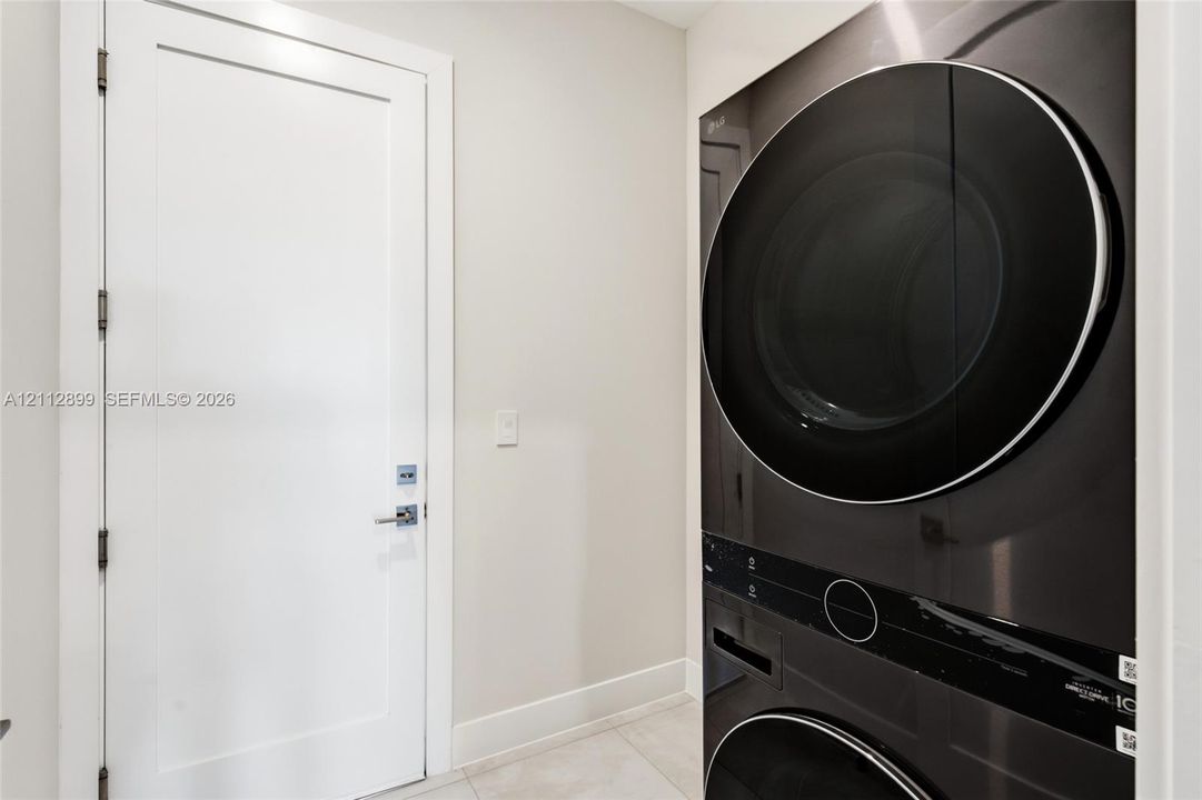 Stacked washer and dryer unit with a sleek black finish, paired with a white Shaker-style door featuring polished chrome hardware and large-format tile flooring
