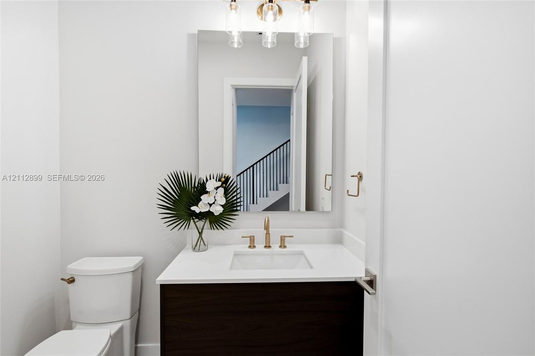 Bathroom featuring a dark wood-finish vanity, white countertop with an integrated basin, wall-mounted rectangular mirror, three-light fixture with cylindrical glass shades, and gold-tone hardware