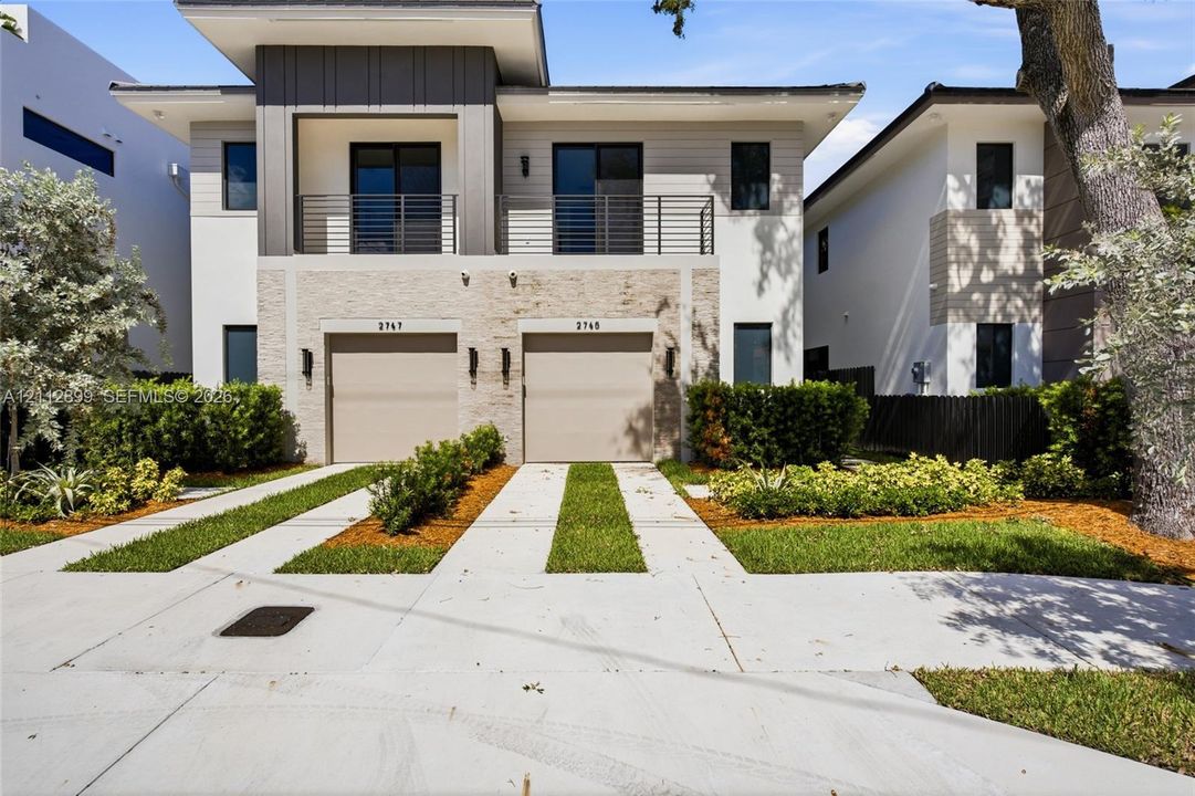 Modern exterior featuring a stacked stone facade, metal-accented second-story balcony with railings, dual two-car garage doors, and concrete paver driveways with integrated grass strips