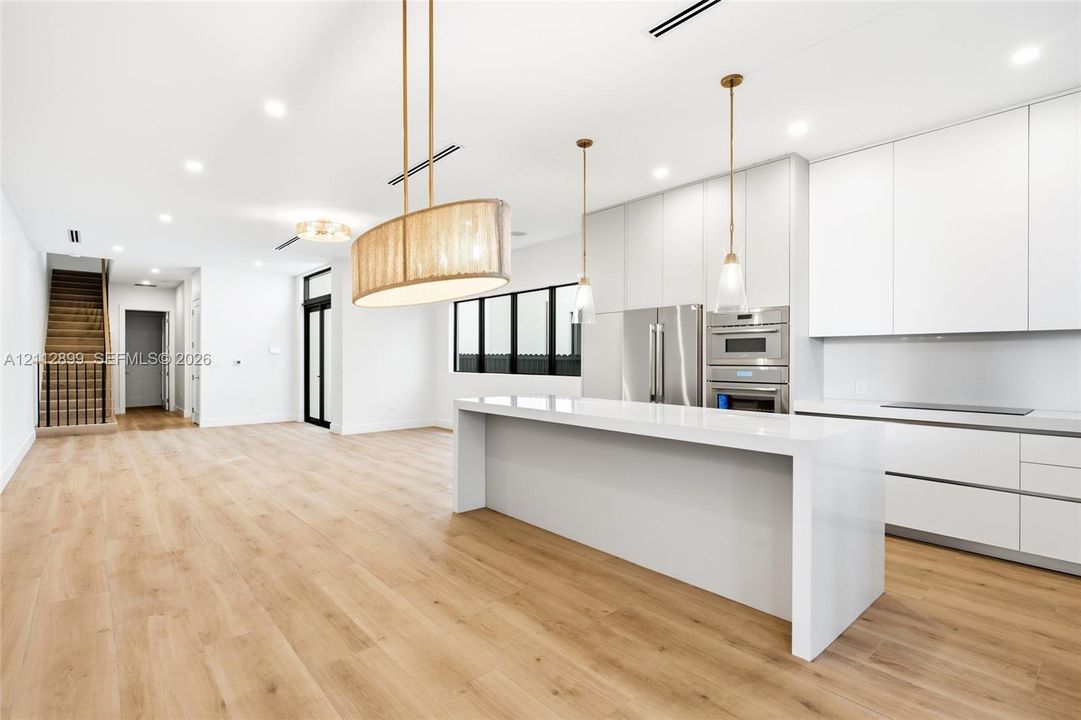 Modern kitchen featuring flat-panel cabinetry, a waterfall-edge quartz island, stainless steel built-in appliances, decorative pendant lighting, and wood-finish flooring