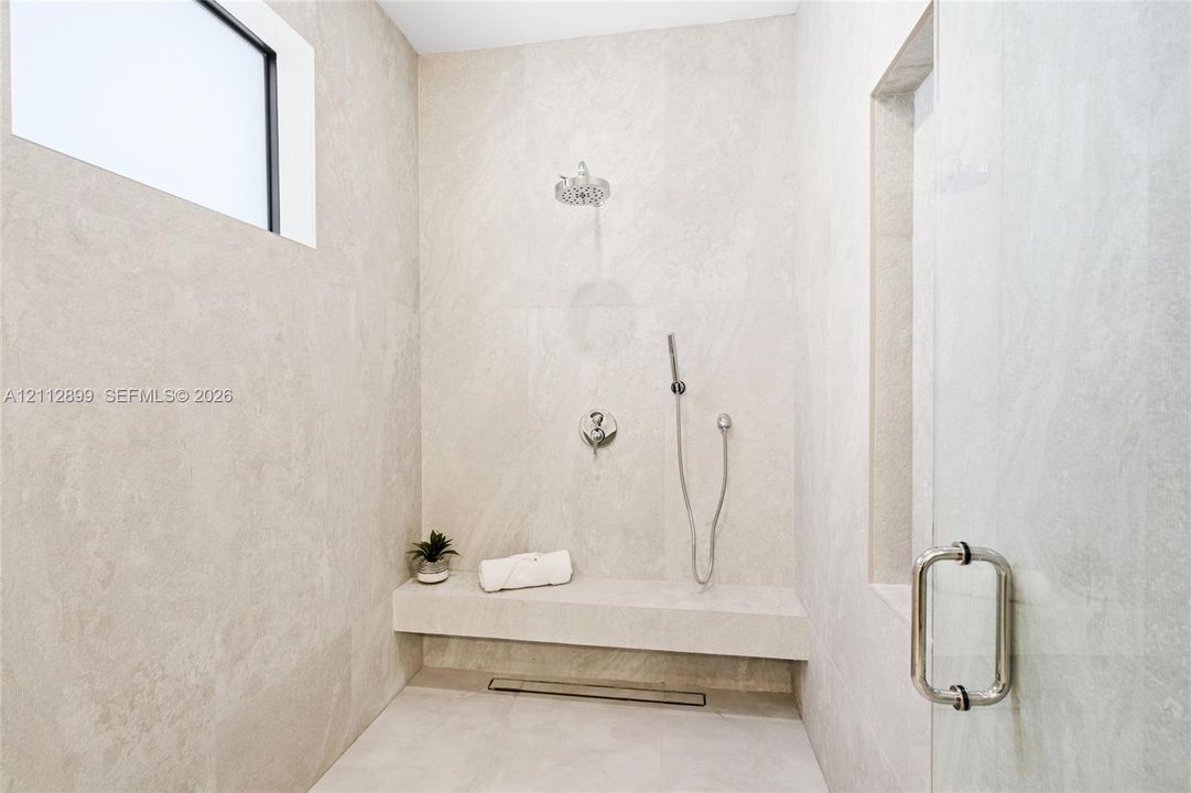 Walk-in shower featuring floor-to-ceiling stone-finish tile, a built-in stone bench, overhead rainfall showerhead, adjustable handheld shower wand, and glass enclosure door