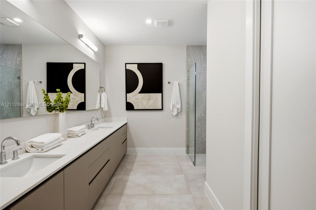 Dual-sink floating vanity with neutral cabinetry, expansive wall-mounted mirror, light-toned stone-finish flooring, and glass-enclosed shower with full-height stone-finish wall cladding
