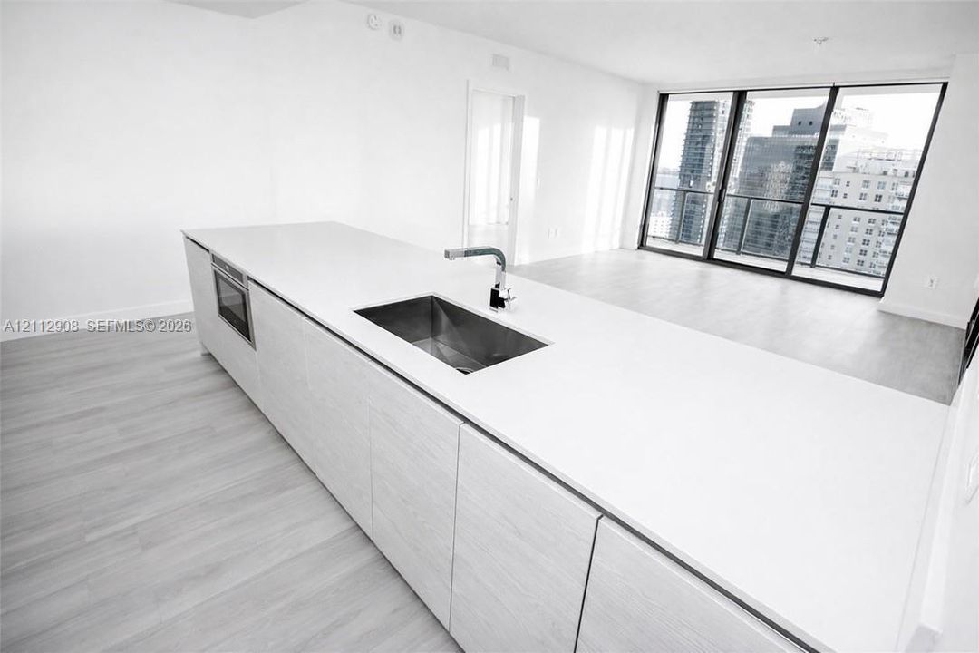Kitchen island featuring light-toned cabinetry, a solid-surface countertop, an integrated stainless steel under-mount sink with a modern faucet, and floor-to-ceiling windows
