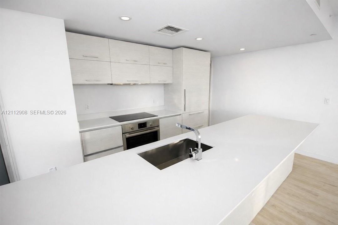 Kitchen featuring flat-panel cabinetry, a quartz countertop island with an undermount stainless steel sink, an electric cooktop, a built-in oven, and wood-finish flooring