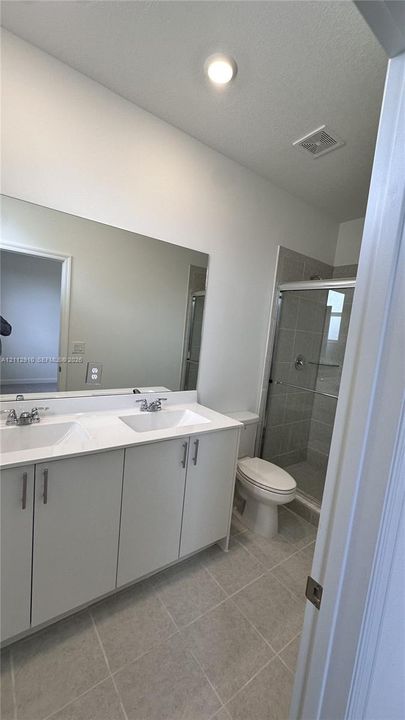 Dual-sink vanity with white cabinetry and solid-surface countertops, walk-in tiled shower with glass enclosure, and neutral tile flooring