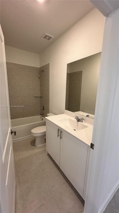 White vanity cabinet with a solid surface countertop, rectangular undermount sink, expansive wall-mounted mirror, ceramic tile flooring, and a tub-shower combination featuring full-height ceramic wall tile