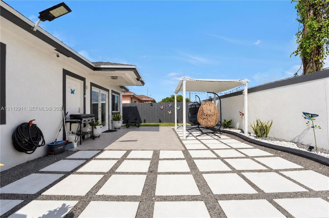 Rear exterior featuring a large concrete paver patio with decorative aggregate inlay, a covered pergola structure, white perimeter masonry walls, and wall-mounted exterior security lighting