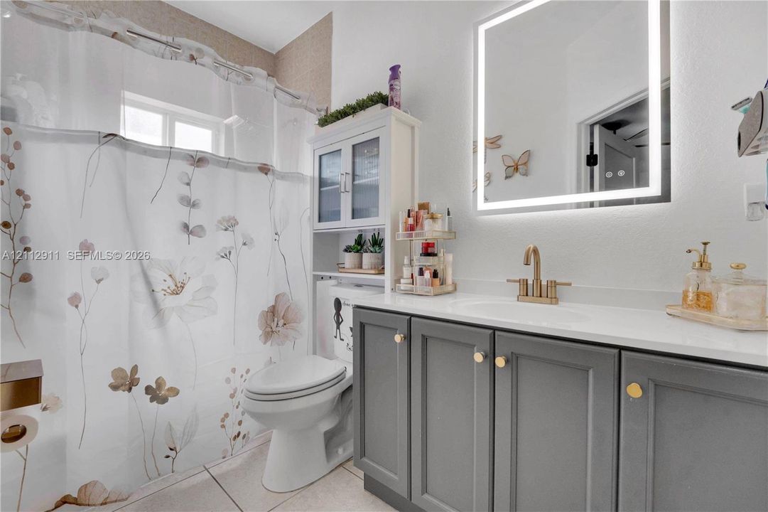 Bathroom featuring a gray shaker-style vanity with a solid-surface countertop, an integrated sink with a brass-finish faucet, and a wall-mounted LED-backlit mirror