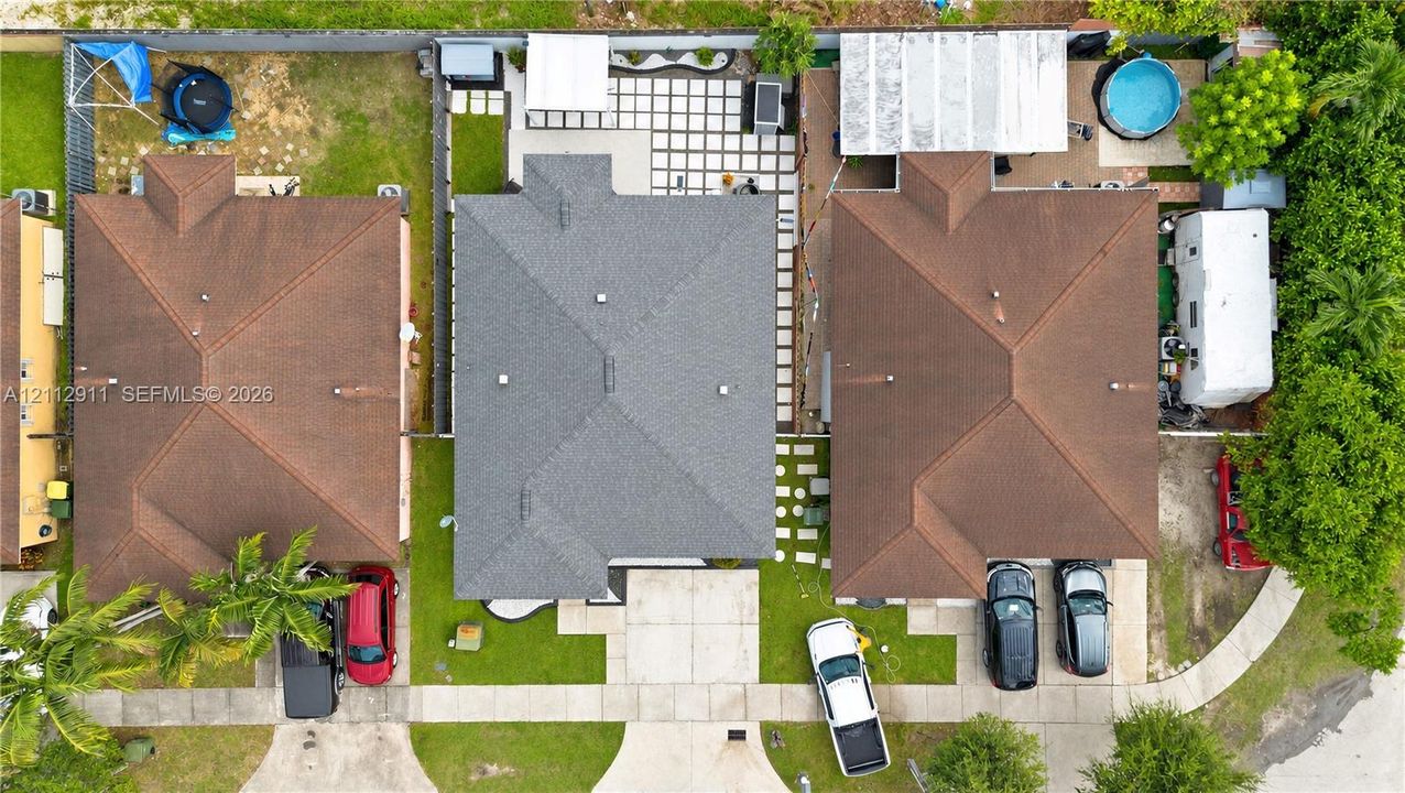Aerial perspective featuring a central residential property with a multi-hipped shingle roof, a concrete driveway, a front lawn, and a rear patio area composed of large-format pavers