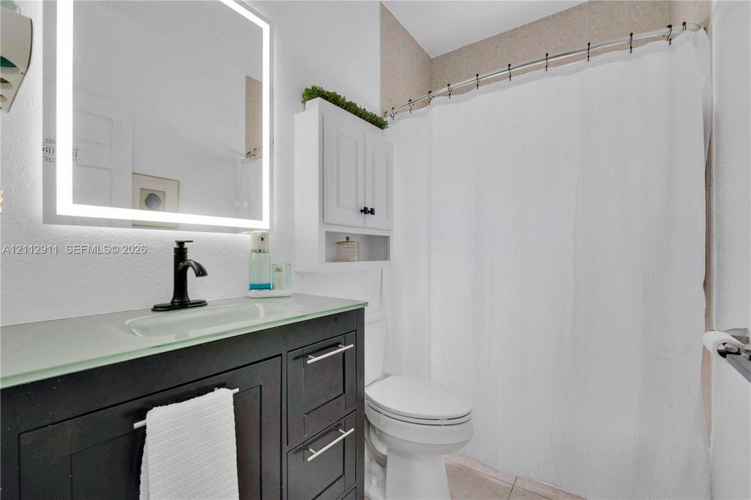 Dark-finish vanity with a glass countertop and integrated sink, matte black faucet, wall-mounted illuminated mirror, and white built-in storage cabinet