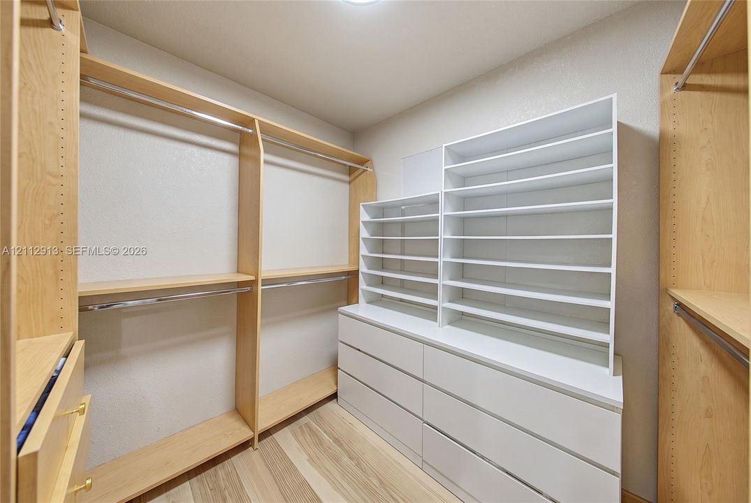 Walk-in closet featuring light wood-finish shelving, custom white cabinetry with integrated drawers, open cubbies, metal garment rods, and wood-finish flooring