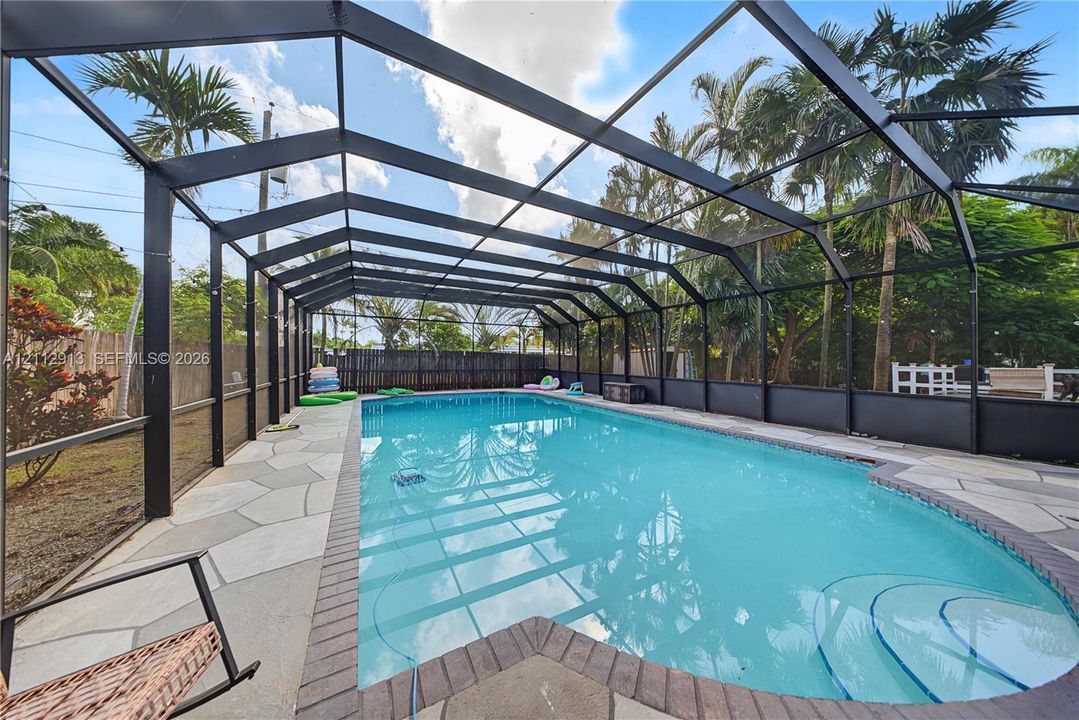 In-ground swimming pool with built-in steps, enclosed by a black metal screened lanai featuring flagstone-style patio pavers and a brick-border finish