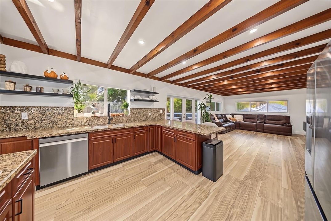 Open-concept kitchen featuring exposed wood ceiling beams, stone countertops, wood-finish flooring, stainless steel appliances, and built-in wall shelving