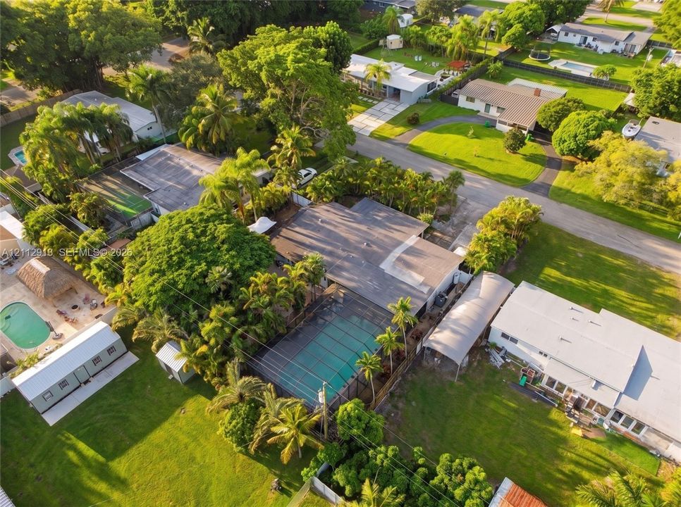 Aerial perspective of a property featuring an in-ground swimming pool with a screened enclosure, a detached metal-roofed carport, mature palm trees, and expansive lawn areas