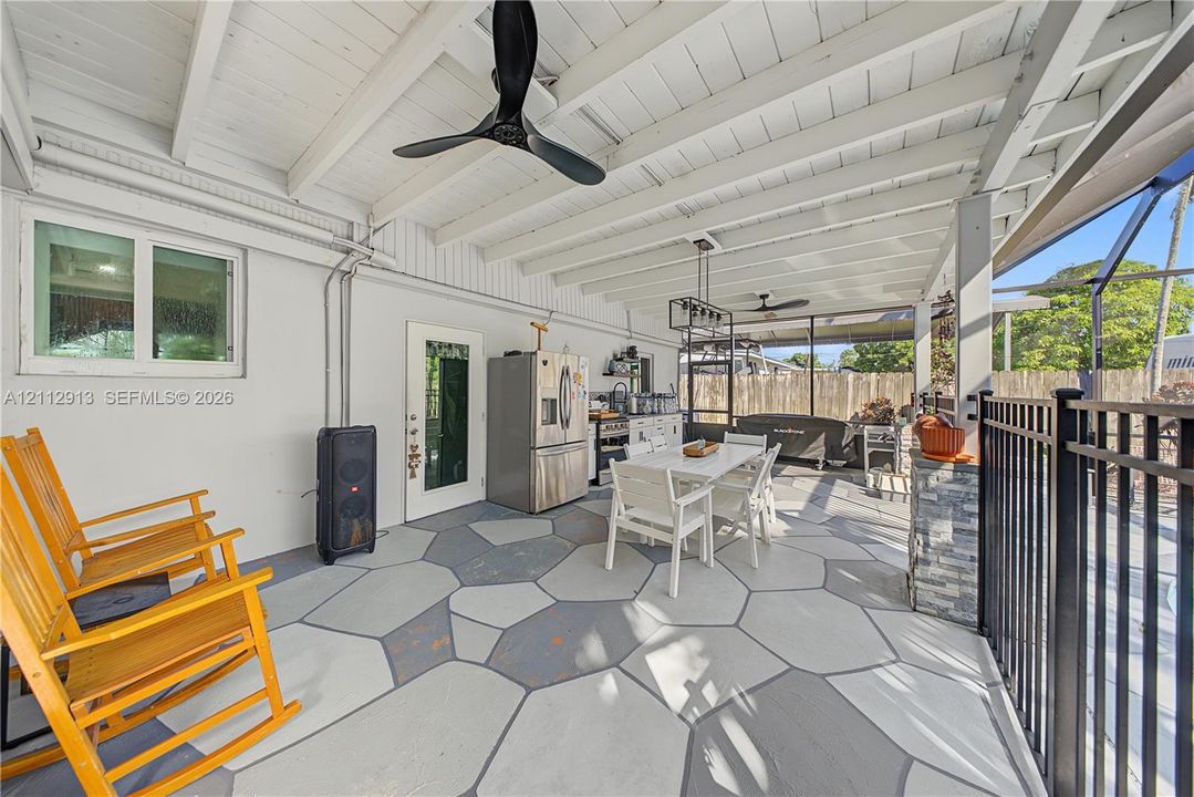 Covered patio featuring a geometric concrete patio surface, exposed beam ceiling, dual matte black ceiling fans, and a stone-accented outdoor counter