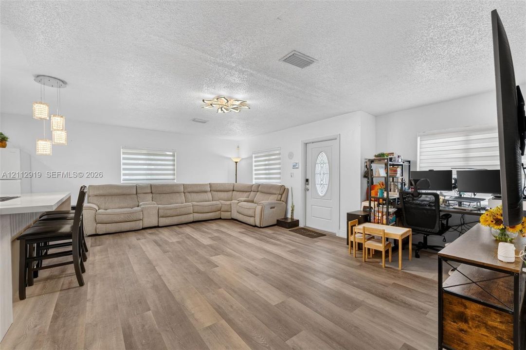 Open-concept space featuring wood-finish flooring, multi-light pendant fixtures, tiered flush-mount ceiling lighting, and windows with integrated horizontal blinds