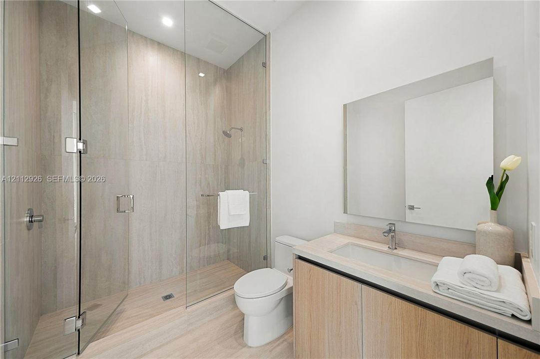 Walk-in shower with floor-to-ceiling tile surround and frameless glass enclosure, wood-finish cabinetry, integrated vanity sink, and wall-mounted mirror