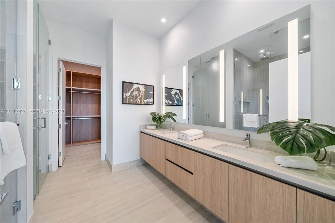 Floating vanity with wood-finish cabinetry, dual integrated sinks, light-embedded mirrors, and large-format tile flooring, featuring a walk-in wardrobe with built-in shelving