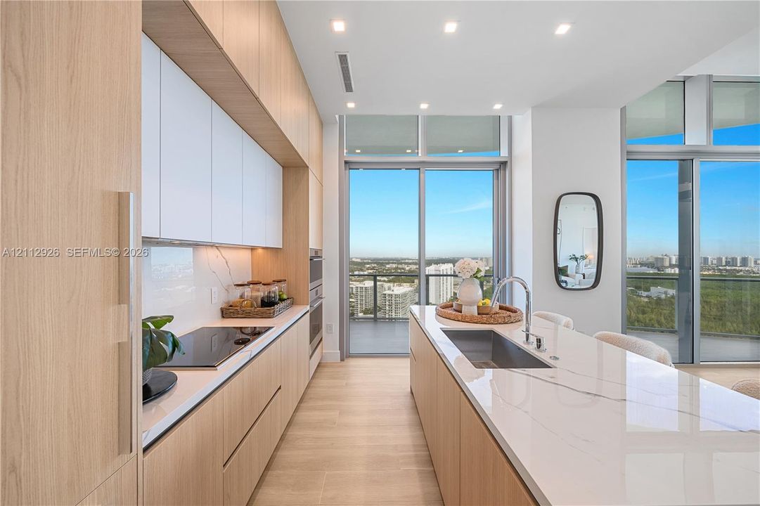 Kitchen featuring floor-to-ceiling wood-finish cabinetry, white matte upper cabinets, stone-surfaced countertops, a center island with an integrated sink, and expansive glass sliding doors