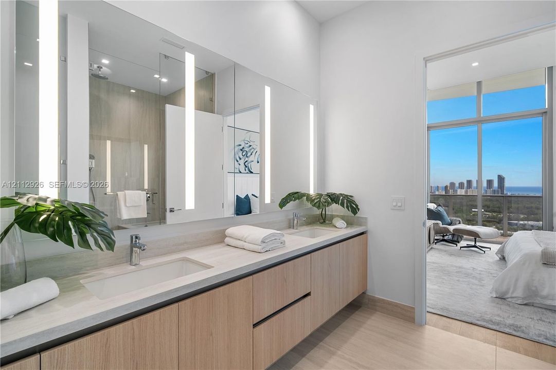 Floating wood-finish vanity with dual integrated rectangular sinks and neutral stone-finish countertops, featuring large-scale mirrors with integrated vertical LED lighting and floor-to-ceiling windows with urban and coastal scenery