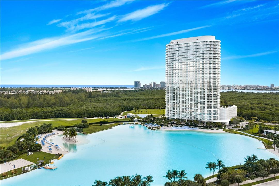 High-rise residential tower featuring extensive balconies, an expansive lagoon, a sandy shoreline, and landscaped green space