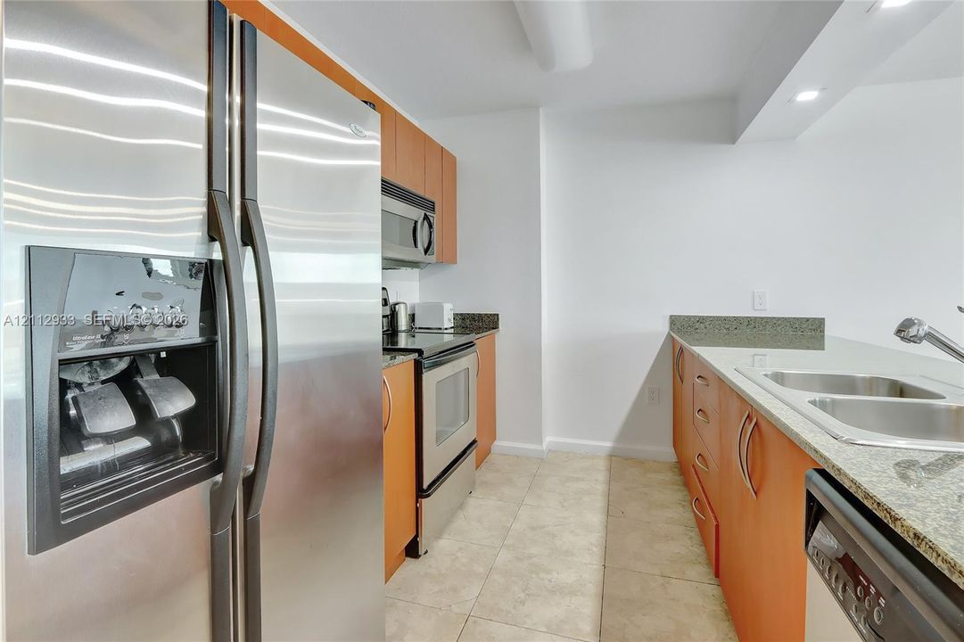 Kitchen featuring stainless steel appliances, wood-finish cabinetry, stone-surface countertops, dual-basin sink, and tile flooring