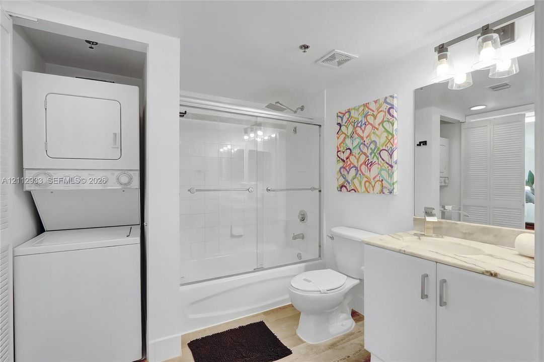 Bathroom featuring a vanity with a stone-top counter, tub with a sliding glass enclosure, integrated laundry closet with a stackable washer and dryer, and three-bulb vanity lighting