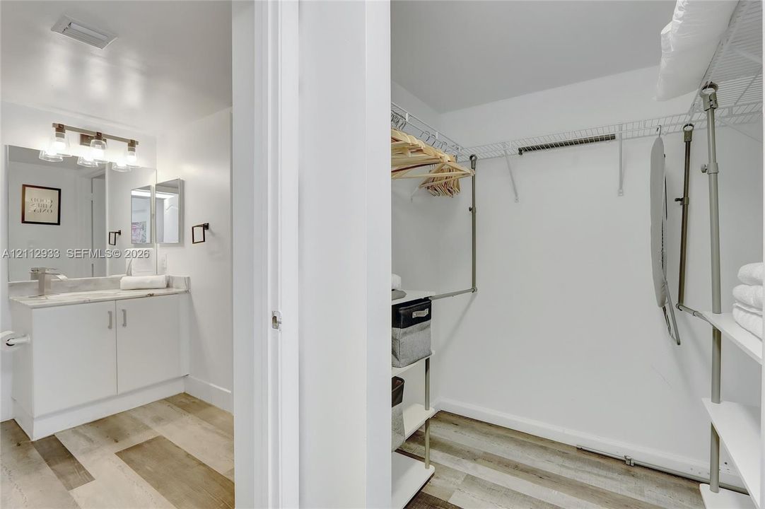 Bathroom vanity featuring a stone countertop, under-mount sink, three-bulb light fixture, and wood-finish flooring, accompanied by a walk-in closet with integrated wire shelving