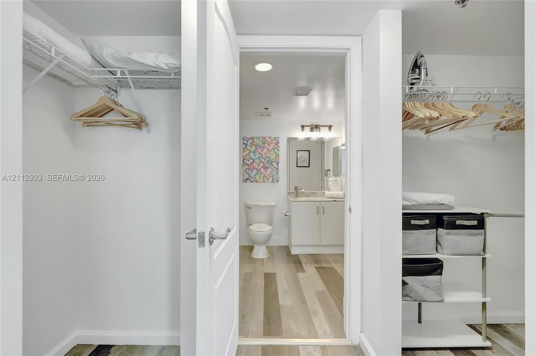 Walk-in closet featuring wire shelving and wood-finish flooring, leading to a bathroom equipped with a vanity, stone-surface countertop, and recessed lighting