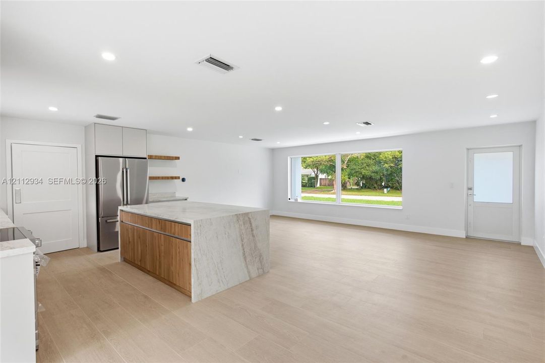 Open-concept kitchen featuring an island with stone-finish waterfall countertops and wood-finish cabinetry, stainless steel refrigerator, recessed lighting, and wood-finish flooring throughout