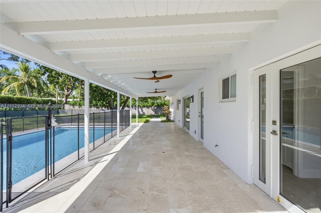 Covered patio featuring stone-tile flooring, beamed ceiling, dual overhead fans, and direct access to an in-ground swimming pool with safety fencing