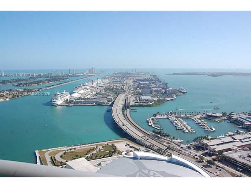 High-elevation aerial perspective featuring expansive coastal waters, an elevated causeway, a harbor with docked vessels, and a surrounding metropolitan cityscape