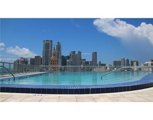 Rooftop swimming pool featuring blue tile coping, stainless steel grab rails, and a glass perimeter railing against a city skyline backdrop