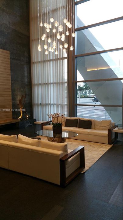 Double-height floor-to-ceiling windows, sculptural multi-pendant chandelier, dark stone flooring, and textured wall paneling
