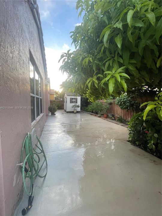 Concrete side yard featuring a perimeter wood fence, mature landscaping, and a detached storage structure