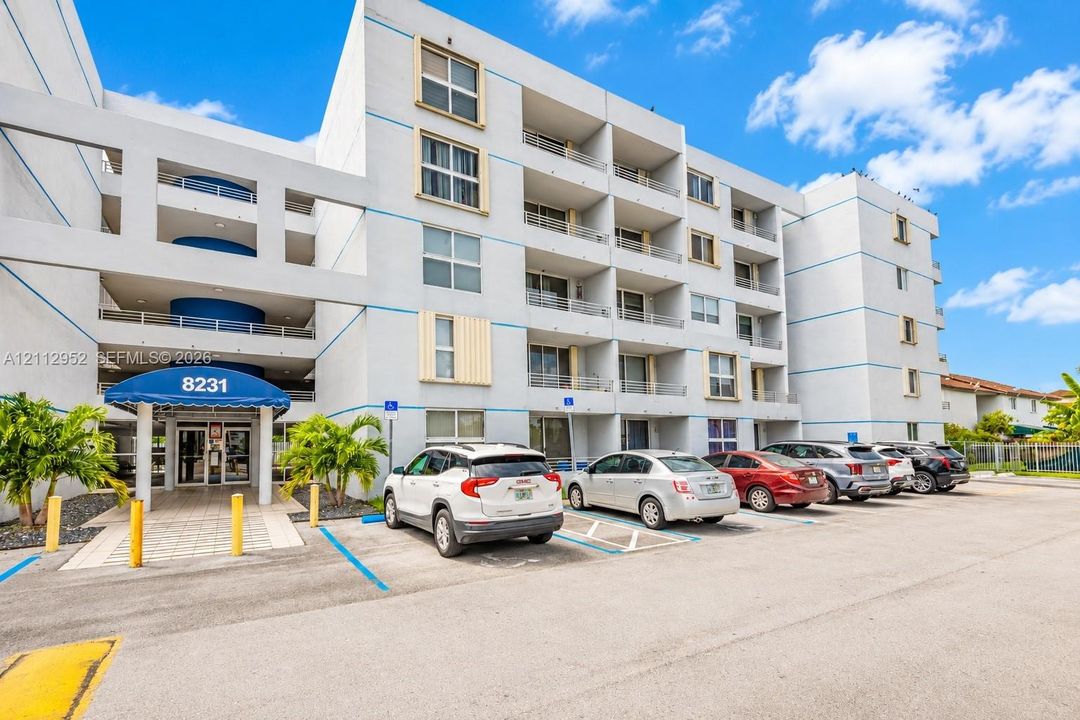 Multi-story stucco exterior featuring recessed balconies with metal railings, a blue fabric entrance canopy, and dedicated surface parking