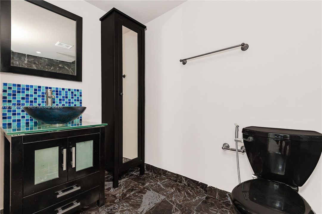 Vanity featuring a dark wood-finish cabinet, glass countertop, and vessel sink with a blue mosaic tile backsplash, complemented by stone-finish flooring and a tall built-in linen cabinet
