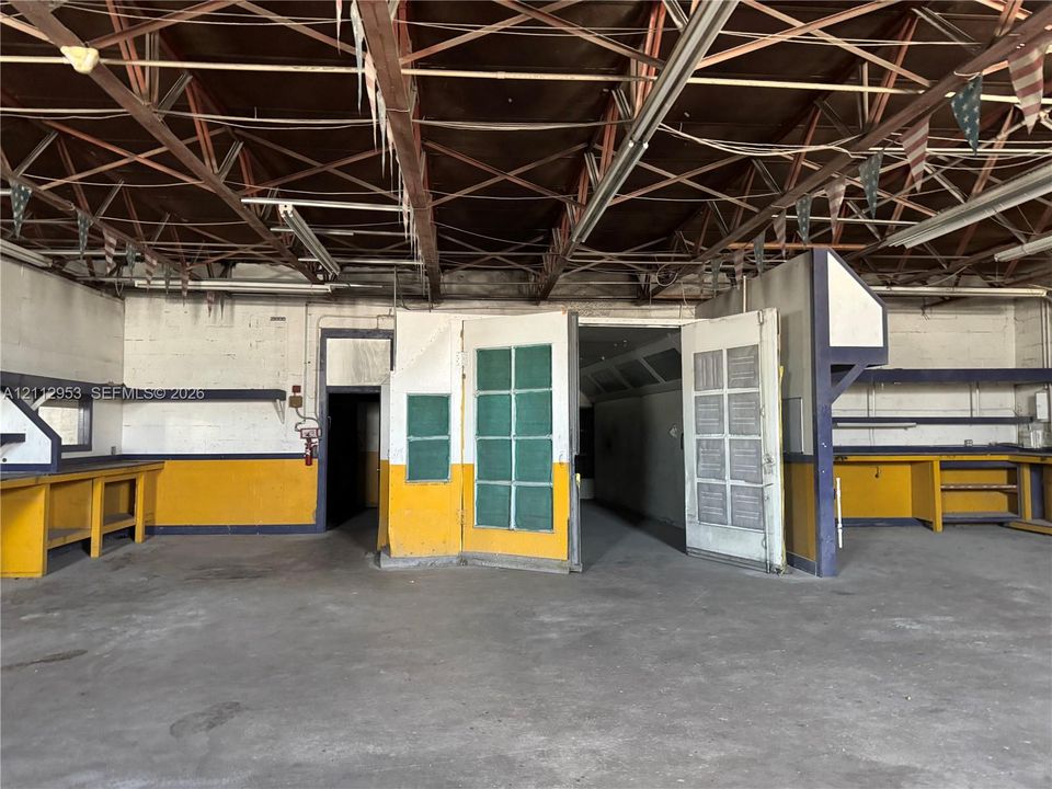 Industrial space featuring exposed steel truss ceiling, concrete flooring, built-in wooden workbenches, and enclosed spray booth area