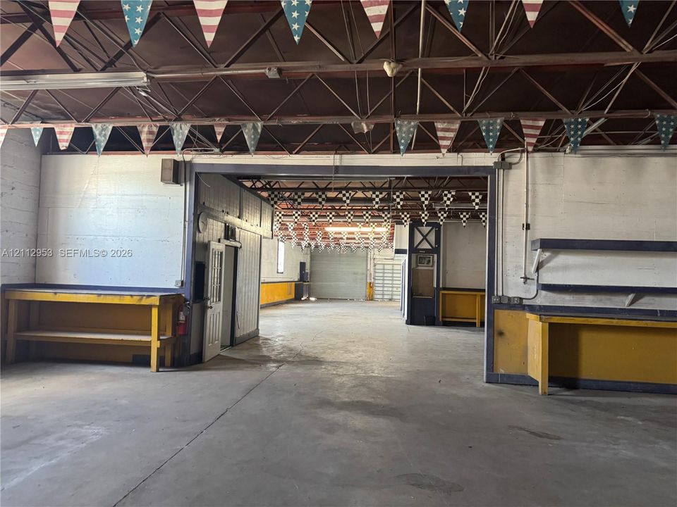 Open-concept industrial workspace featuring exposed metal roof trusses, concrete flooring, built-in wooden work counters, and a roll-up bay door