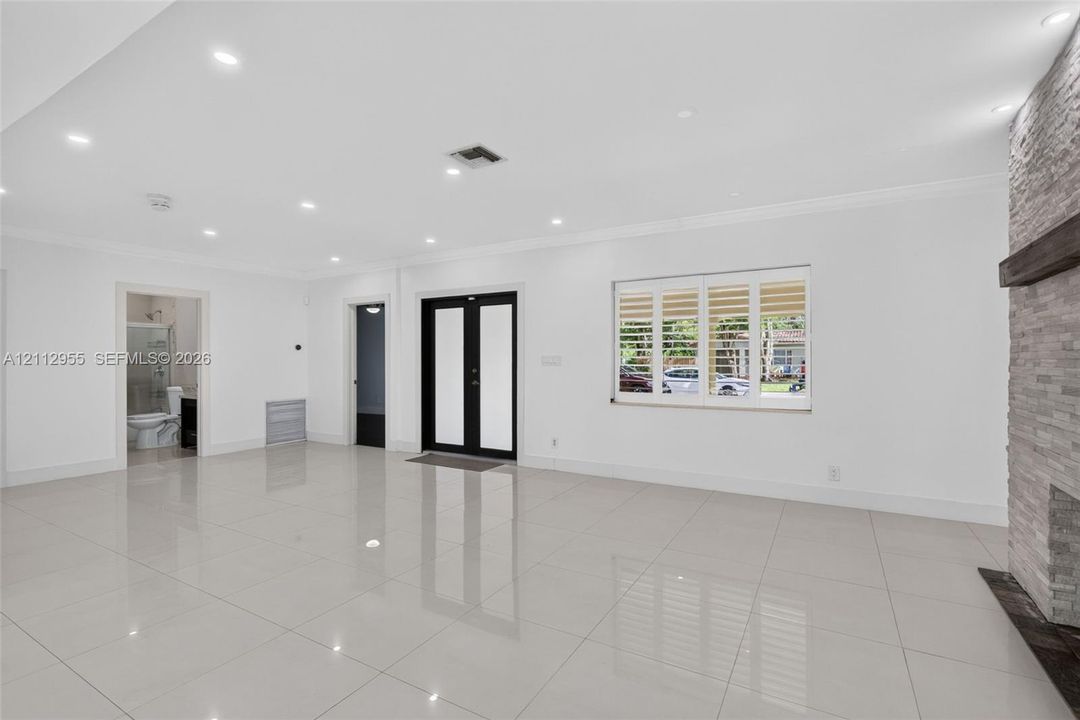 Large-format tile flooring, recessed ceiling lighting, stone-clad fireplace with a timber mantel, double-entry front door with glass insets, and windows featuring plantation shutters